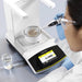 Elevated view of a laboratory technician wearing safety glasses and protective gloves using an electronic pipette to dispense liquid into a vessel on a Sartorius Secura 120 analytical balance displaying an active digital readout.