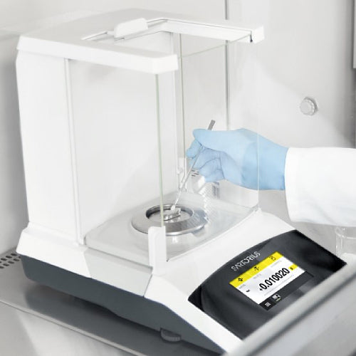 A gloved laboratory technician using a micro-spatula to dispense a sample onto the weighing pan inside the glass draft shield chamber of a Sartorius Secura 120 analytical balance showing an active digital touchscreen display.