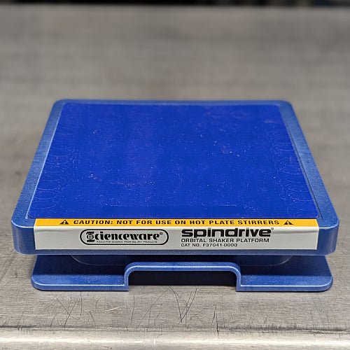 Straight-on close-up studio view of the blue Scienceware Spindrive orbital shaker platform resting on a stainless steel surface, highlighting the front label with branding, model name, catalog number, and caution warning