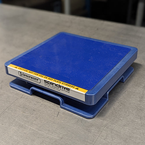 Three-quarter side-angled studio view of the blue Scienceware Spindrive orbital shaker platform resting on a metallic surface, displaying the front warning label and side-profile cutouts of the molded base