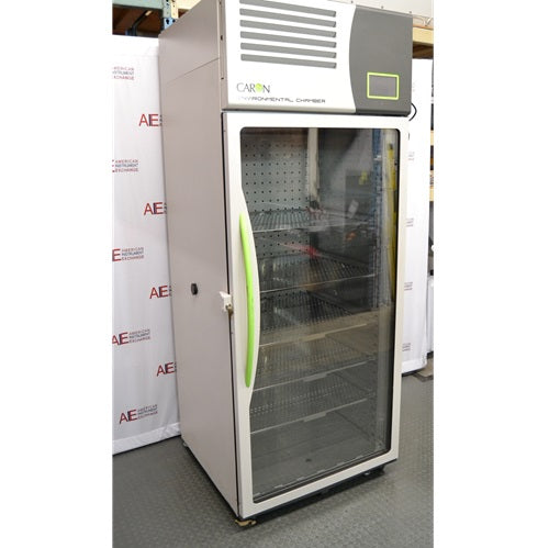 A side-angled view of the tall, upright Caron 7000-33-1 environmental chamber, showcasing the unit's side profile, full-length glass door with a bright green handle, and top-mounted control interface, all positioned in front of an American Instrument Exchange backdrop.
