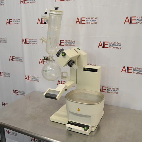 An angled side-front view of a Buchi R-300 rotary evaporator system resting on a stainless steel laboratory benchtop in front of an American Instrument Exchange backdrop, displaying the heating bath, lift arm, drive unit, glass condenser assembly, and attached receiving flask.