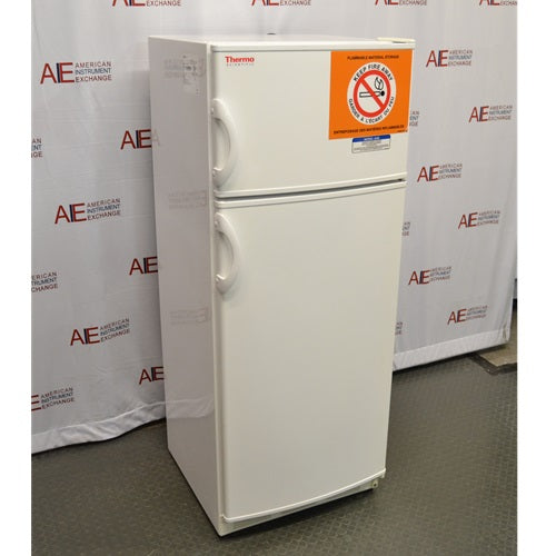 An angled side view of a white Thermo Scientific 10FCEETSA flammable material combination refrigerator/freezer on a lab floor, showing the top freezer compartment with a flammable storage safety label, the main refrigerator compartment below, and side door handles against an American Instrument Exchange backdrop.