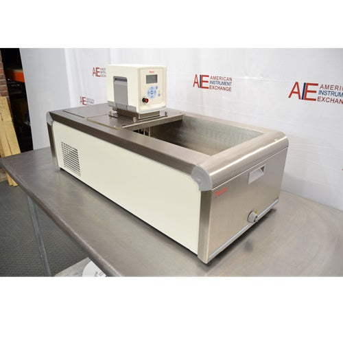 A high-angled side perspective view of a Thermo Scientific A10B refrigerated bath circulator sitting on a metal workbench in front of an American Instrument Exchange backdrop, displaying its cream side panel with ventilation grille, stainless steel front with drain valve, open bath basin, and top-mounted digital control head.