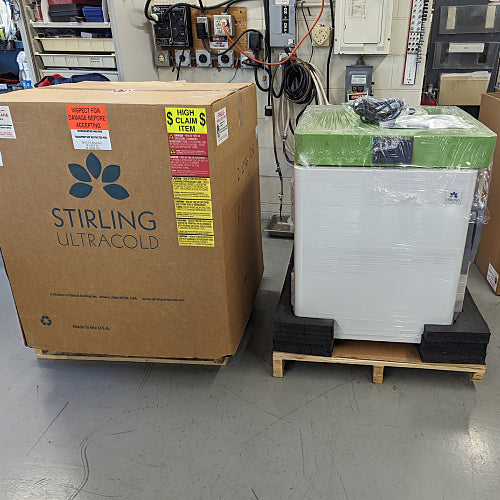 Warehouse view of a Stirling Ultracold SU105UE freezer next to its branded cardboard shipping carton on wooden pallets.