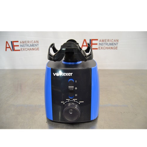 A straight-on front view of a blue and black Heathrow Scientific Vortexer featuring a top tube holder attachment, central power switch, speed control knob, and American Instrument Exchange background logo.

