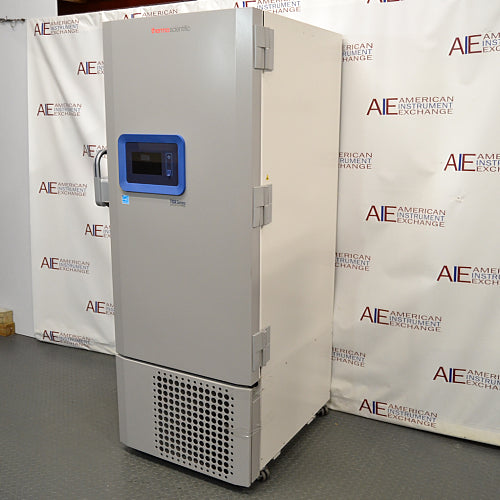 Straight-on studio view of a Thermo Scientific TSX40086 upright ultralow temperature freezer standing on a grey floor against a branded backdrop, featuring a solid grey outer door, ergonomic handle, integrated blue digital touchscreen interface, TSX Series branding, and a lower perforated ventilation panel