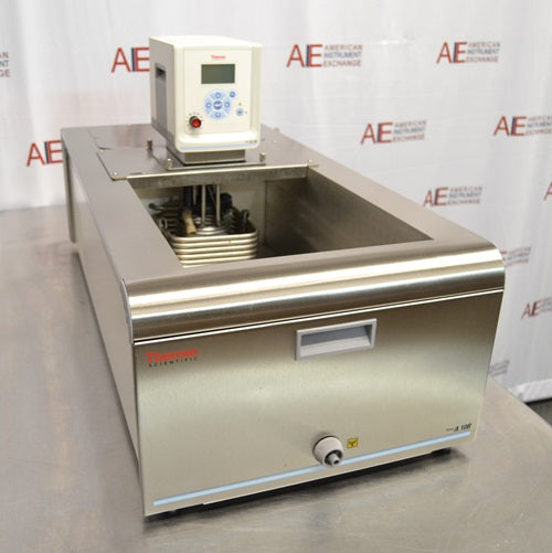 A top-angled front view of a Thermo Scientific A10B refrigerated bath circulator resting on a metal surface against an American Instrument Exchange backdrop, showing the open bath chamber with internal cooling coils, top-mounted digital control unit, stainless steel front casing, drain valve, and model logo.