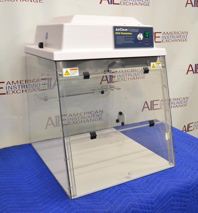 Front angled perspective view of an AirClean Systems AC624LFUV PCR workstation resting on a blue cloth-covered surface, highlighting its clear acrylic walls, front folding sash, and upper control console housing