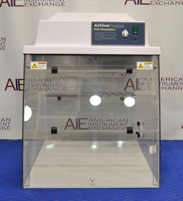 Straight-on front view of an AirClean Systems AC624LFUV PCR workstation with clear acrylic panels, overhead control console, and white interior base resting on a blue surface