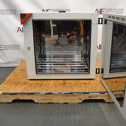 Front view of a Binder FD-S 115 mechanical convection oven with its side-hinged door swung open, showing the polished stainless steel interior chamber, adjustable wire shelves, and a reflective back panel