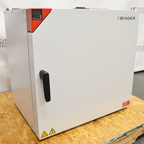 Three-quarter angle product view of a Binder FD-S 115 mechanical convection oven, emphasizing its white cabinet structure, red corner control interface, and door handle resting on a wooden pallet base