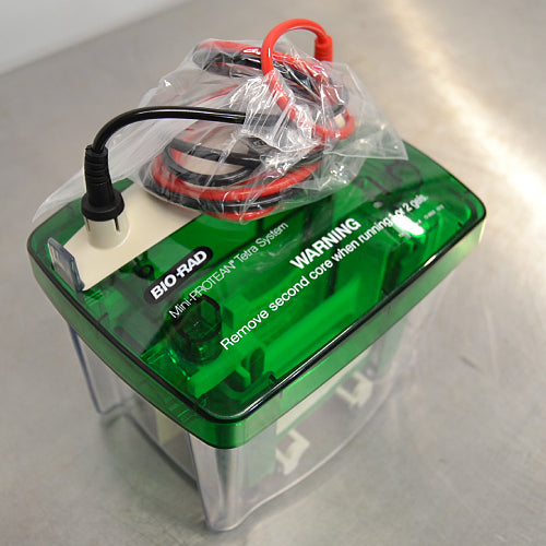 Left angle view of a Bio-Rad Mini-PROTEAN Tetra Cell electrophoresis system, showing the green safety lid with warning text and the attached power cables