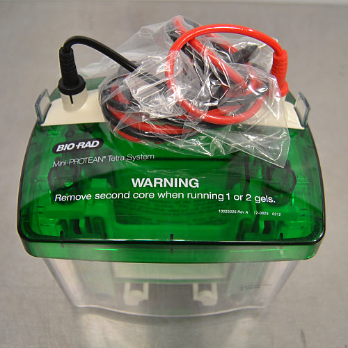 Top angle view of a Bio-Rad Mini-PROTEAN Tetra Cell electrophoresis system, featuring a green safety lid with warning text regarding core removal, along with coiled black and red power cables resting on top