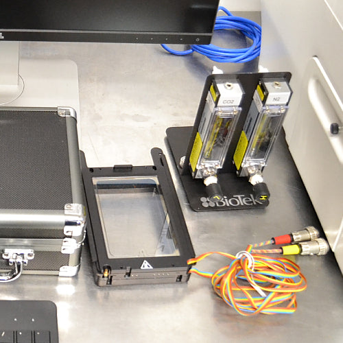 Close-up view of accessories for a BioTek Cytation 5 cell imaging multimode reader on a stainless steel surface, featuring dual CO2 and N2 gas flow meters on a stand, a specialized stage adapter insert, and a wired sensor harness