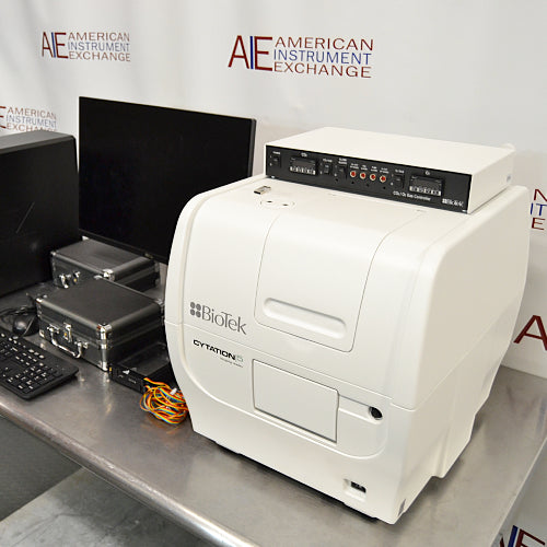 Front angled perspective view of a BioTek Cytation 5 cell imaging multimode reader with a top-mounted gas controller, positioned on a stainless steel work surface next to a computer monitor, keyboard, and accessory cases