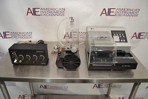 Front eye-level view of a BioTek ELx405UV Select microplate washer system resting on a stainless steel table, complete with its accompanying valve module, vacuum pump, and fluid bottle setup