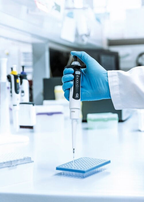 Lab researcher in blue gloved hand using BRANDTECH Transferpette pro single-channel pipette to dispense liquid into a 96-well plate in a laboratory setting