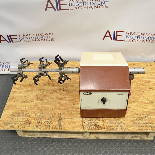 Elevated front product view of a Burrell wrist action laboratory shaker, featuring its brown and beige housing, front control dial, and horizontal shaker arm with attached flask clamps resting on a wooden pallet
