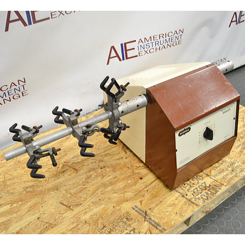 Left three-quarter angle product view of a Burrell wrist action laboratory shaker, featuring its brown and beige housing, speed control dial, and horizontal arm with multiple flask clamps on a wooden pallet