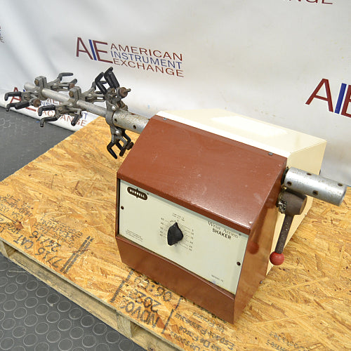 Right three-quarter angle product view of a Burrell wrist action laboratory shaker, showing its brown and beige housing, front control panel, side extension arm, and flask clamp assembly resting on a wooden pallet