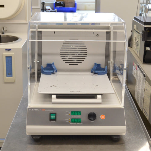 Front product view of a Corning LSE shaking incubator model 6790, featuring its clear acrylic housing hood, internal platform with blue flask clamps, rear ventilation grille, and front control panel with green digital LED displays and rotary knob