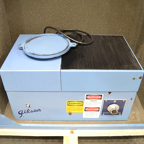 Front product view of a Gilson sieve shaker housed inside a sound enclosure, showcasing its blue housing, brand logo, safety and danger labels, and control dial on a wooden pallet base