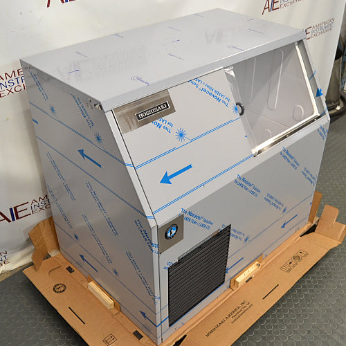 Front angled perspective view of a new-in-box Hoshizaki F300BAF flaker ice maker on a wooden pallet, showing its stainless steel body wrapped in protective film, lower front vent grille, and side panel
