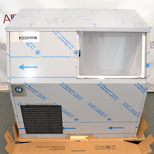 Front eye-level view of a new-in-box Hoshizaki F300BAF flaker ice maker resting on a wooden pallet, featuring stainless steel housing covered in protective blue-printed film, a lower front louvered vent, and an open bin door