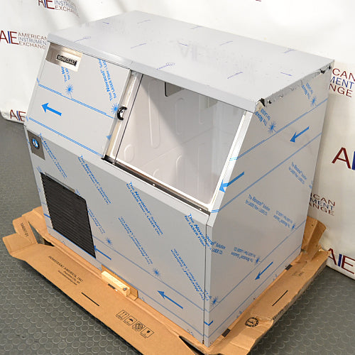 Right angled perspective view of a new-in-box Hoshizaki F300BAF flaker ice maker on a pallet, highlighting its stainless steel body encased in protective shipping film, open top bin door, and lower front vent