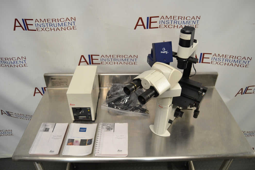Overhead angled view of a Leica DMIL inverted microscope with fluorescence setup on a stainless steel table, displaying the microscope unit, power supply box, bundled cables, instruction manuals, and camera attachment