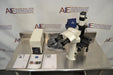 Overhead angled view of a Leica DMIL inverted microscope with fluorescence setup on a stainless steel table, displaying the microscope unit, power supply box, bundled cables, instruction manuals, and camera attachment