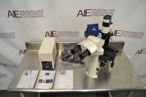 Overhead angled view of a Leica DMIL inverted microscope with fluorescence setup on a stainless steel table, displaying the microscope unit, power supply box, bundled cables, instruction manuals, and camera attachment