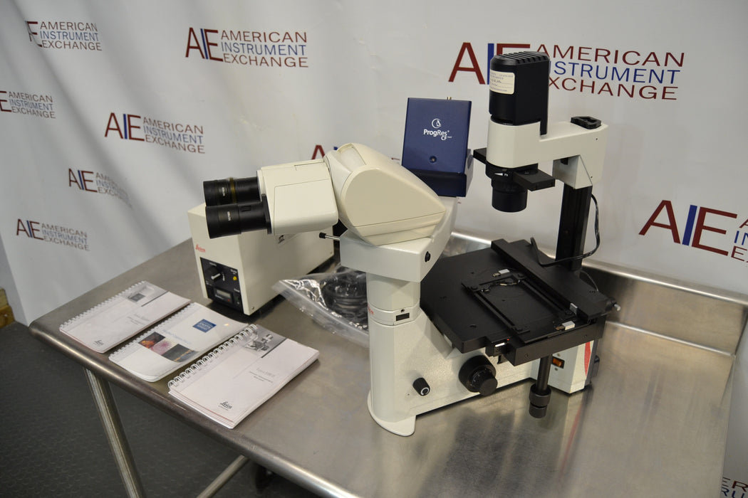 Angled perspective view of a Leica DMIL inverted microscope with fluorescence system on a stainless steel workstation, featuring the main optical body, binocular eyepieces, mechanical stage, camera equipment, power unit, and operation manuals