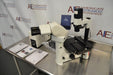 Angled perspective view of a Leica DMIL inverted microscope with fluorescence system on a stainless steel workstation, featuring the main optical body, binocular eyepieces, mechanical stage, camera equipment, power unit, and operation manuals