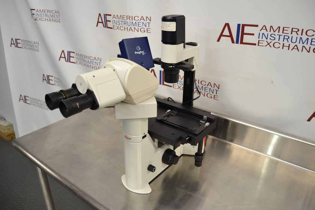Close-up side profile view of a Leica DMIL inverted microscope with fluorescence system resting on a stainless steel table, highlighting the main vertical stand, binocular viewing head, mechanical stage, and attached digital camera unit