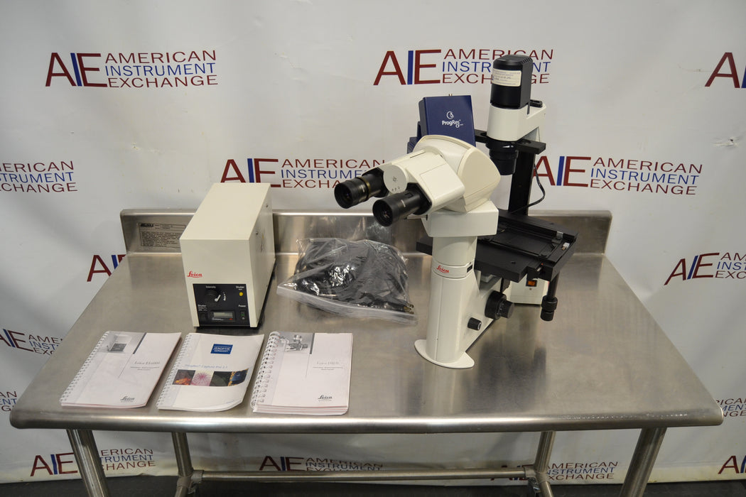 Front-facing view of a Leica DMIL inverted microscope with fluorescence setup arranged on a stainless steel table, including the main microscope body with binocular head, a power supply unit, manuals, a bag of cables, and an attached camera component