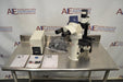 Front-facing view of a Leica DMIL inverted microscope with fluorescence setup arranged on a stainless steel table, including the main microscope body with binocular head, a power supply unit, manuals, a bag of cables, and an attached camera component