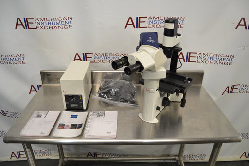 Front-facing view of a Leica DMIL inverted microscope with fluorescence setup arranged on a stainless steel table, including the main microscope body with binocular head, a power supply unit, manuals, a bag of cables, and an attached camera component