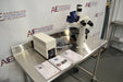 Left angled perspective view of a Leica DMIL inverted microscope with fluorescence system set up on a stainless steel table, showing the power supply unit, manuals, packaged cables, and main microscope body with optical head