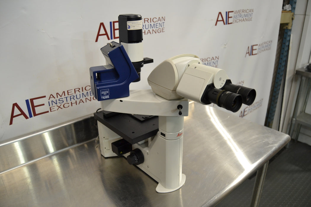 Right angled perspective view of a Leica DMIL inverted microscope with fluorescence attachment on a stainless steel work table, emphasizing the side profile, optical head, digital camera apparatus, and mechanical stage