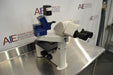 Right angled perspective view of a Leica DMIL inverted microscope with fluorescence attachment on a stainless steel work table, emphasizing the side profile, optical head, digital camera apparatus, and mechanical stage