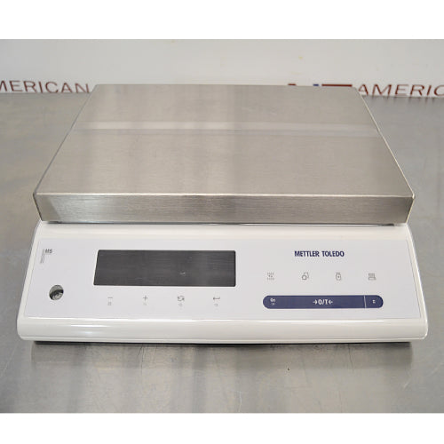 Front product view of a Mettler Toledo MS12001L/03 high-precision toploading balance, featuring a large rectangular stainless steel weighing pan, white housing, digital display screen, and integrated control keypad on a stainless steel lab bench