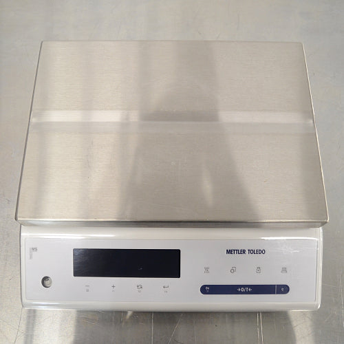 Top-down product view of a Mettler Toledo MS12001L/03 high-precision toploading balance, featuring a large flat stainless steel weighing platter, white base housing, digital display screen, and control panel on a stainless steel surface