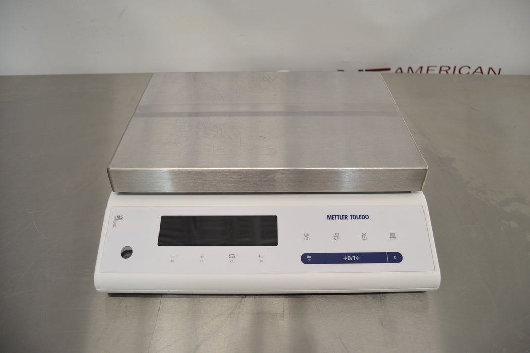 Front eye-level view of a Mettler Toledo MS32001L precision balance resting on a stainless steel surface, showcasing its large stainless steel weighing pan and digital display control panel