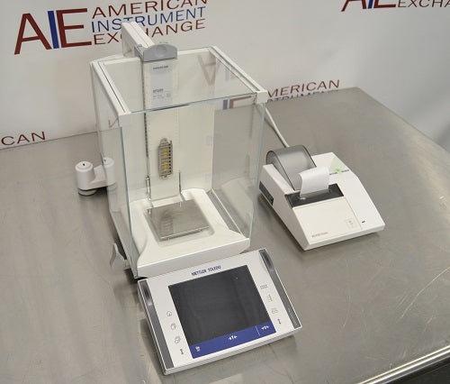 Angled perspective view of a Mettler Toledo XP205 analytical balance on a stainless steel surface, featuring its glass draft shield, front touchscreen terminal, interior weighing pan, and a connected laboratory printer