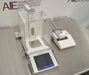 Angled perspective view of a Mettler Toledo XP205 analytical balance on a stainless steel surface, featuring its glass draft shield, front touchscreen terminal, interior weighing pan, and a connected laboratory printer