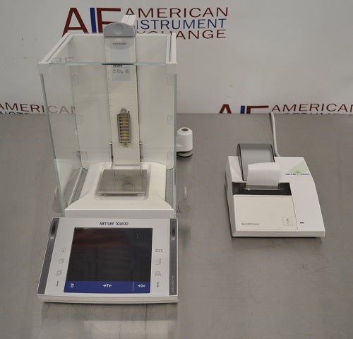 Front high-angle view of a Mettler Toledo XP205 analytical balance on a stainless steel counter, featuring its glass draft shield, separate touchscreen terminal, weighing pan, and a connected laboratory printer