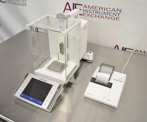 Top-down view of a Mettler Toledo XP205 analytical balance resting on a stainless steel counter, featuring its protective glass draft shield, front touchscreen interface, internal weighing pan, and a connected label printer