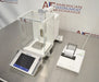 Top-down view of a Mettler Toledo XP205 analytical balance resting on a stainless steel counter, featuring its protective glass draft shield, front touchscreen interface, internal weighing pan, and a connected label printer